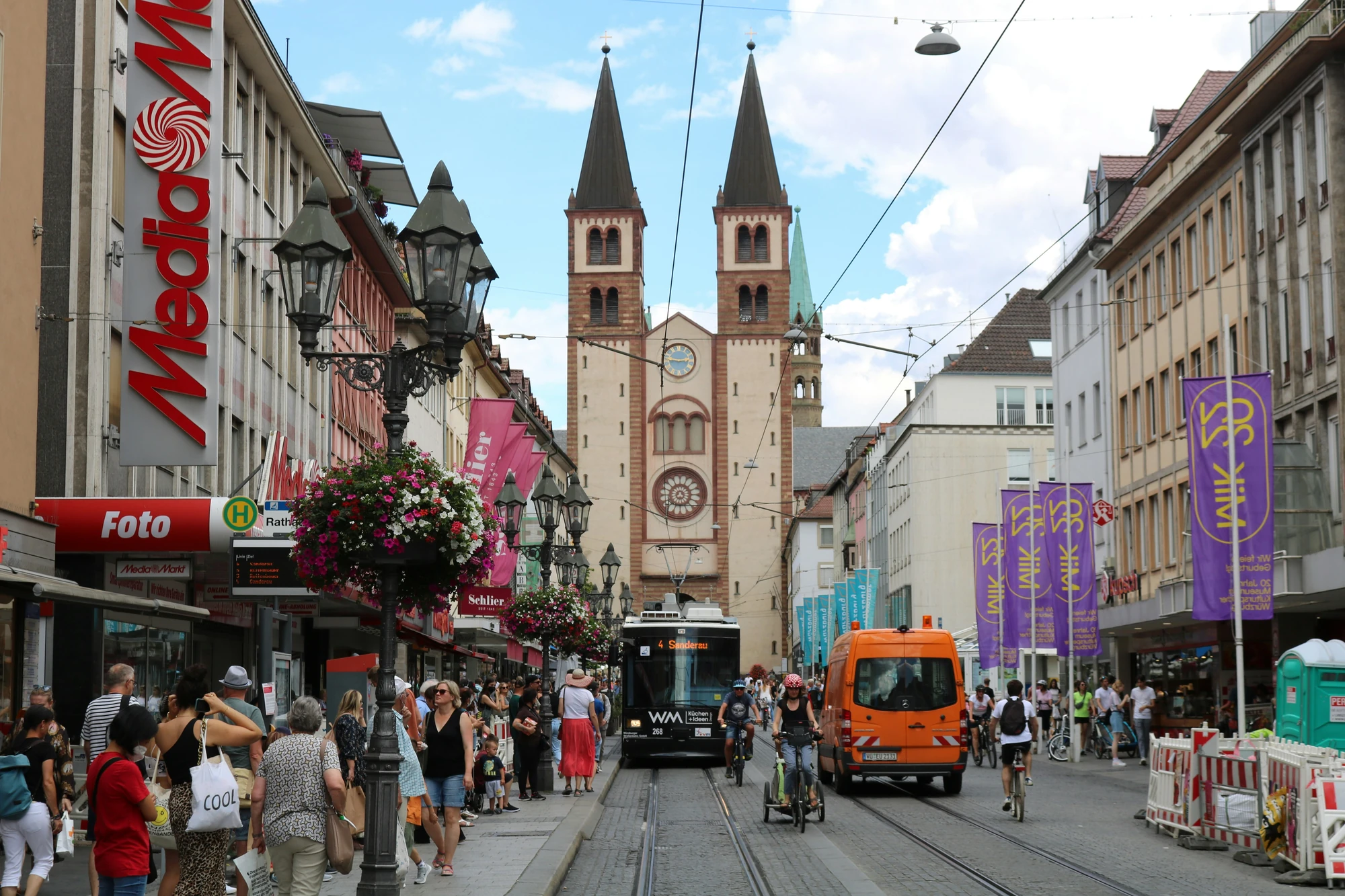 Belebte Einkaufsstraße in Würzburg, im Hintergrund ragen die Türme des Kiliansdoms empor. Eine Straßenbahn fährt entlang der Menschenmenge, die das Stadtleben genießt.