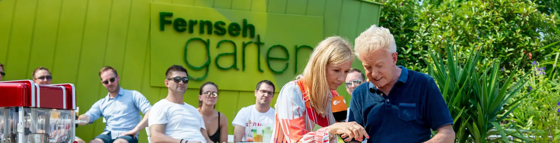 Menschen sitzen und Andrea Kiewel sowie Armin Roßmeier stehen im Freien vor einer grünen Wand mit dem Schriftzug 'Fernsehgarten' in der TV Show ZDF Fernsehgarten (im Bannerformat)