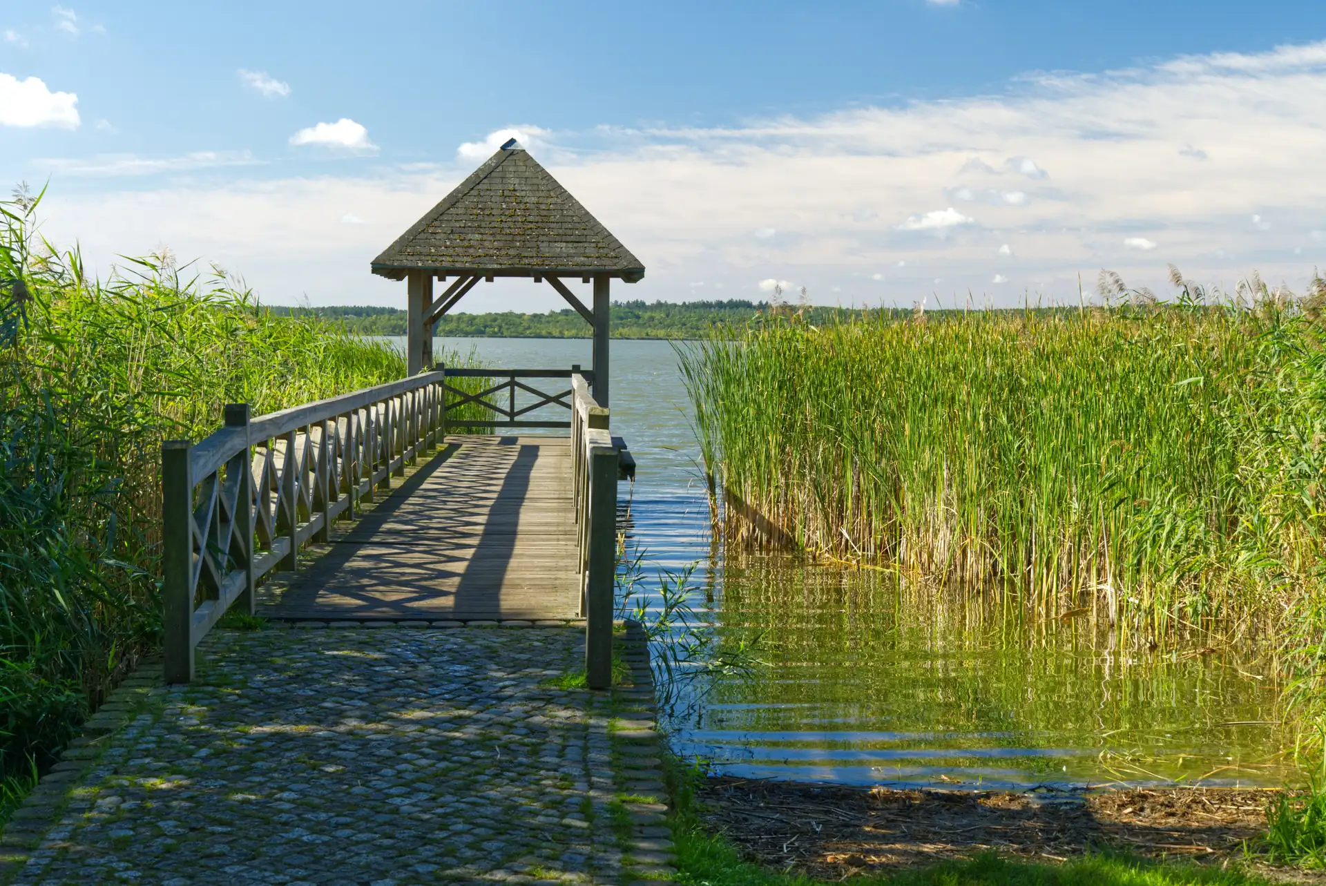 Steg mit Holzhütte am Ufer des Zierker Sees umgeben von Schilf und Wasser unter blauem Himmel mit Wolken