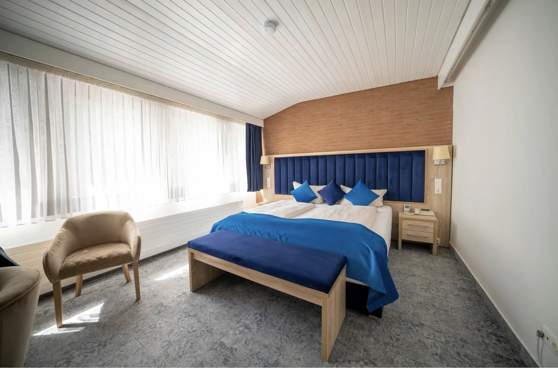 Doppelzimmer im Hotel Zur Heidschnucke mit großem Bett, blauer Bettdecke und Kissen, Holzbank am Fußende, zwei Sesseln und großen Fenstern mit weißen Vorhängen
