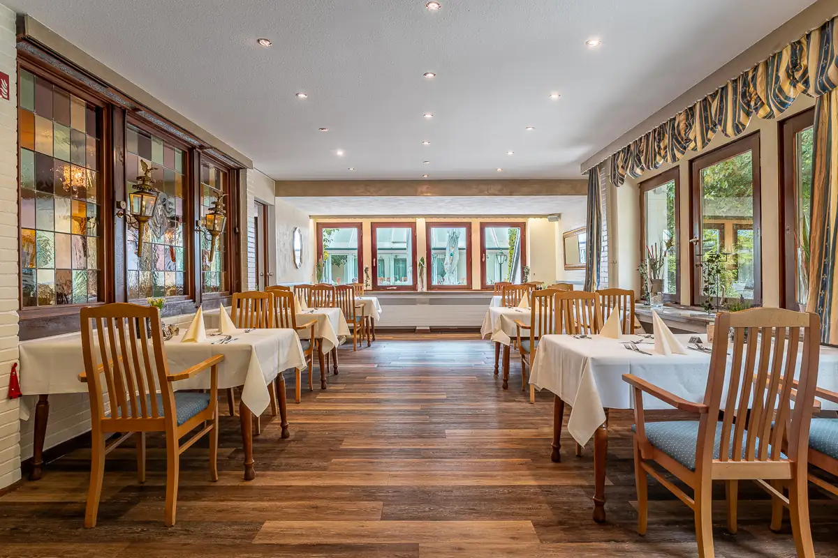 Helles Restaurant mit Holzboden, weißen Tischdecken auf Tischen und Holzstühlen, dekorativen Fenstern und Vorhängen im Hotel Zur Heidschnucke
