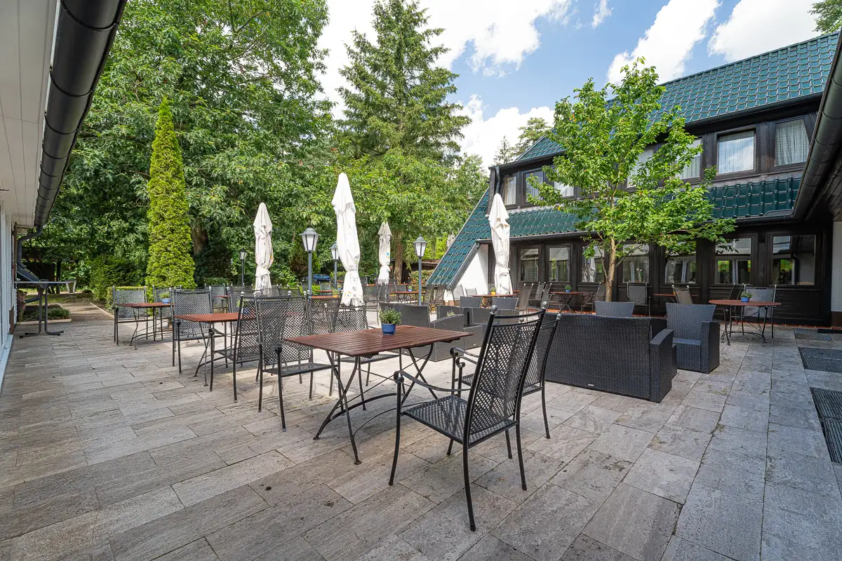 Terrasse des Hotels Zur Heidschnucke mit mehreren Tischen, Stühlen und geschlossenen Sonnenschirmen vor einem zweistöckigen Gebäude und umgeben von Bäumen