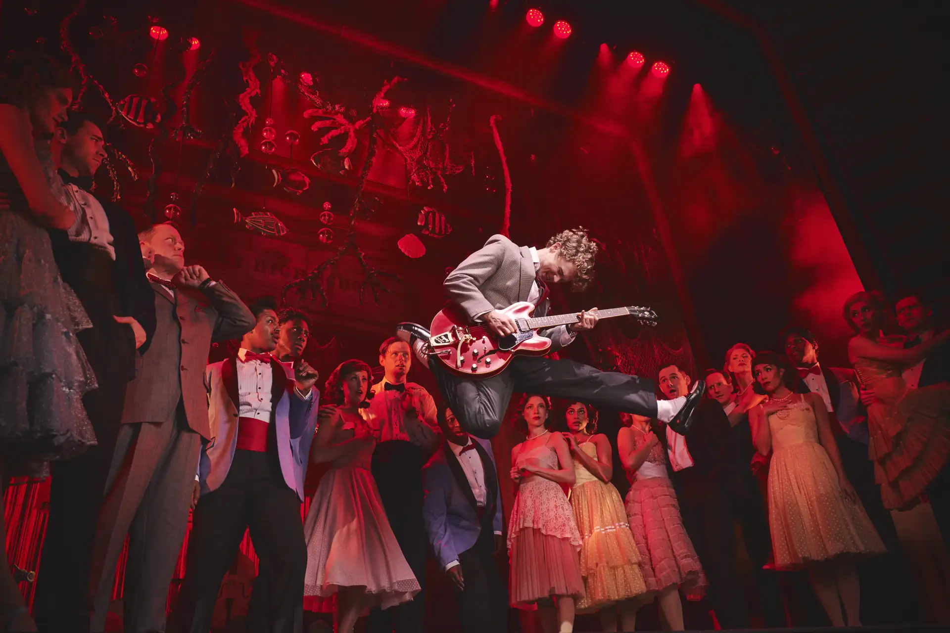 Zurück in die Zukunft – Das Musical, Szenenmotiv: Marty McFly (Caden Brauch) springt mit Gitarre auf Bühne, umgeben von tanzenden Menschen in festlicher Kleidung, rotes Bühnenlicht