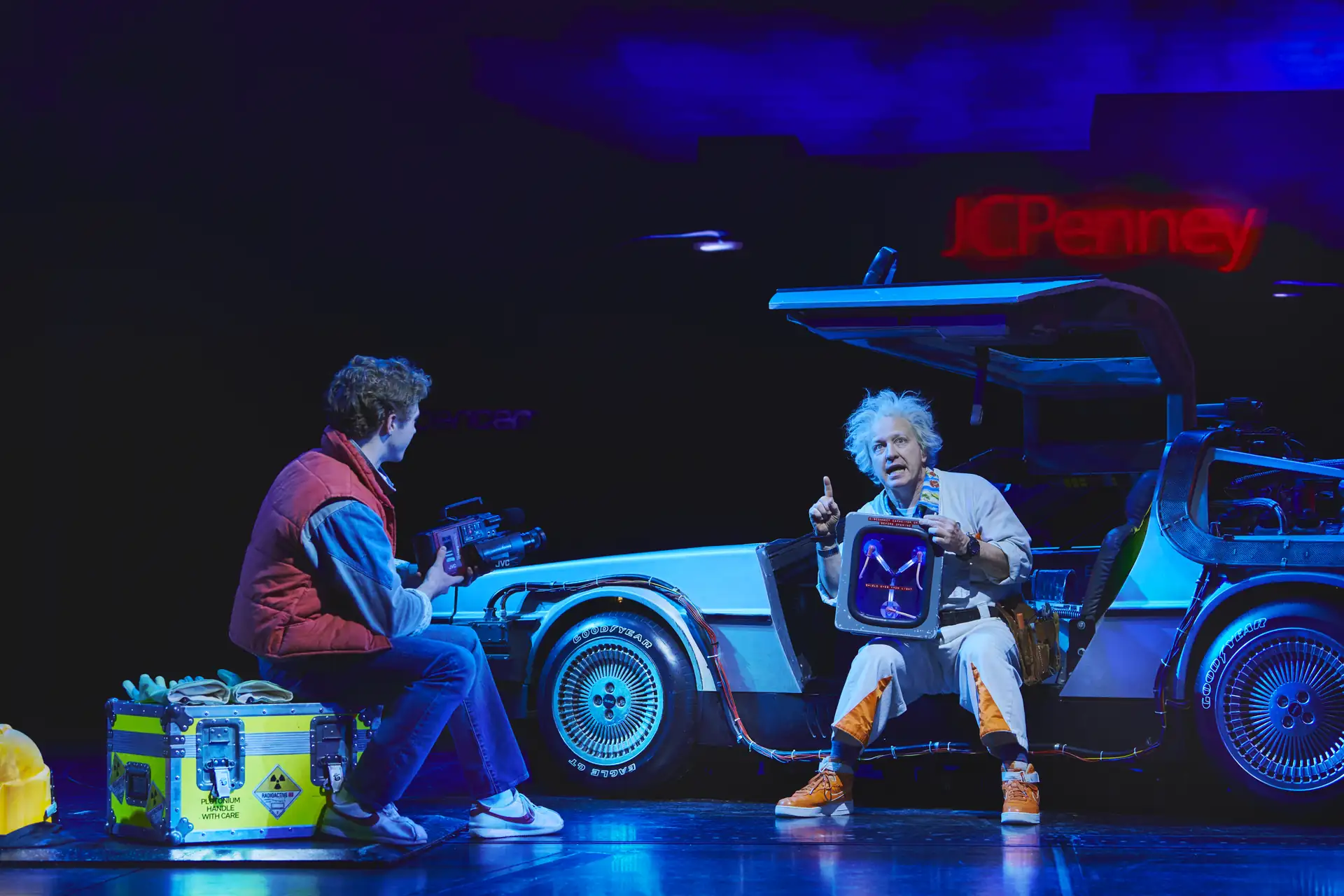 Zurück in die Zukunft – Das Musical, Szenenmotiv: Marty McFly (Caden Brauch) & Doc Brown (Cory english) mit einem DeLorean-Auto, einer sitzt auf einem Koffer, der andere hält ein Gerät mit einem leuchtenden Bildschirm.