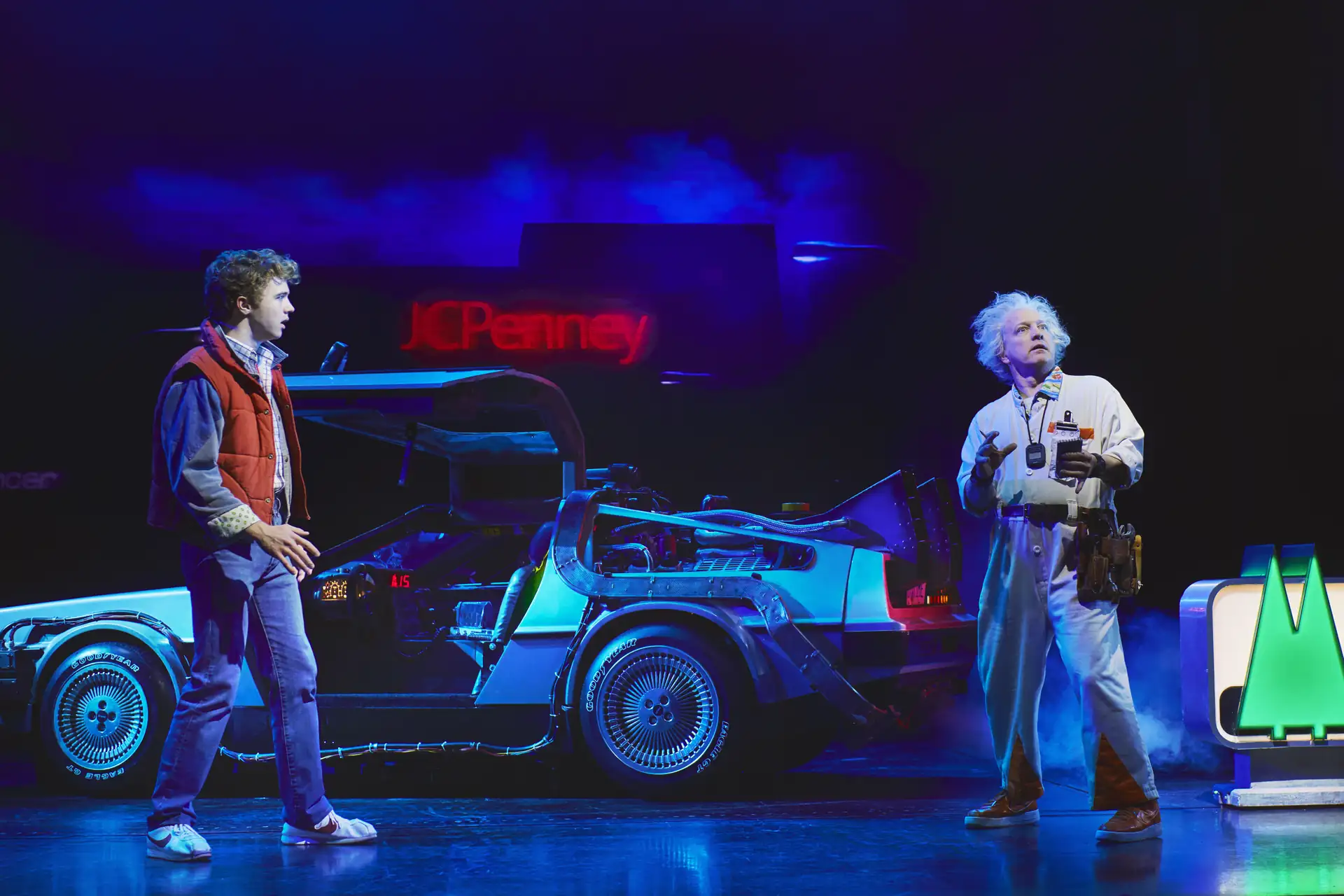 Zurück in die Zukunft – Das Musical, Szenenmotiv: Marty McFly (Caden Brauch) & Doc Brown (Cory english) neben einem silbernen DeLorean-Auto. Marty trägt eine rote Weste, Brown einen weißen Overall mit technischen Geräten.
