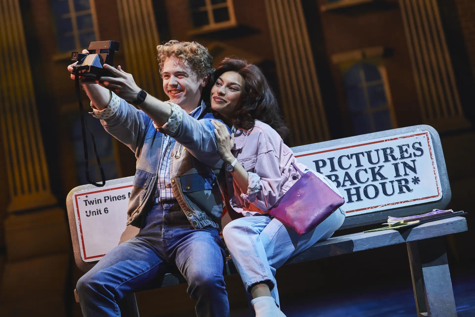 Zurück in die Zukunft – Das Musical, Szenenmotiv: Marty McFly (Caden Brauch) & Jennifer Parker (Talia Palamathanan) sitzen auf einer Bank mit dem Schild 'Pictures Back in Hour' und macht ein Selfie mit einer Sofortbildkamera