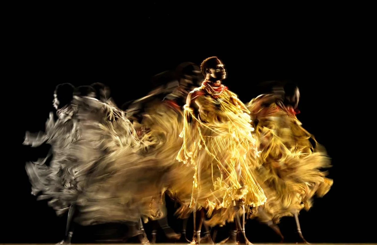 Das Bild zeigt eine dynamische und künstlerische Darstellung eines Tänzers, der in einer verschwommenen Bewegung eingefangen wurde. Die langbeinigen und wehenden Stoffe, die der Tänzer trägt, scheinen im Tanz zu verschwimmen, was eine faszinierende Darstellung der Bewegung und Energie schafft. Der Hintergrund ist dunkel, was den Fokus vollständig auf die Tanzbewegung und die lebendige Farbenpracht lenkt, die durch die fließende Kleidung und den Tanzstil erzeugt wird. Diese Art von Darstellung könnte in einer Ausstellung wie 